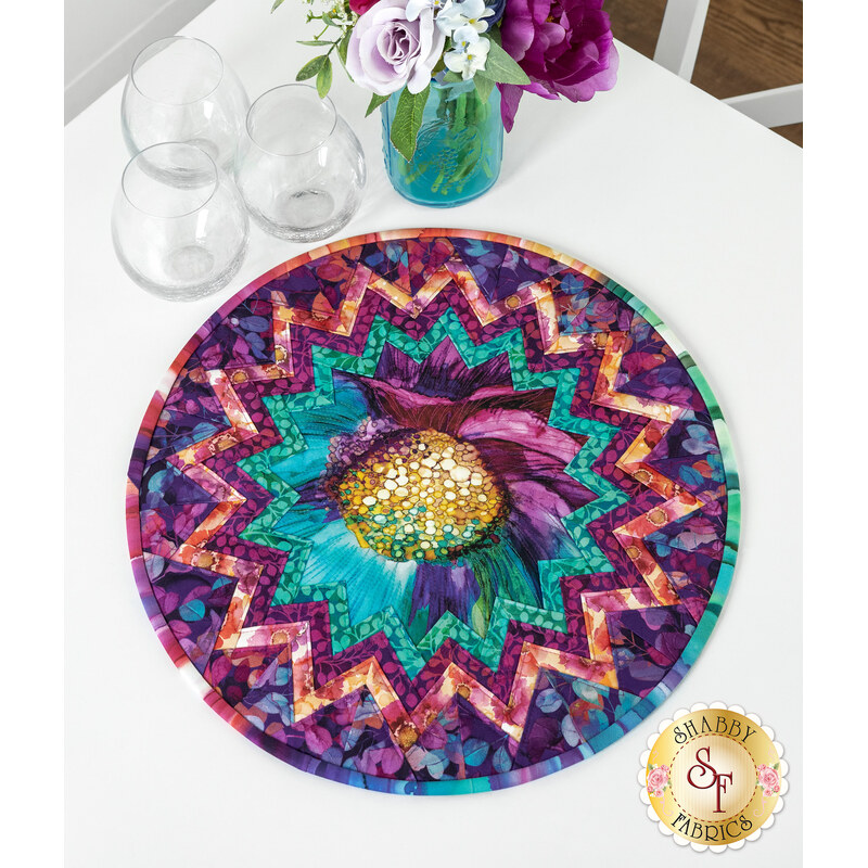 The completed Point of View Folded Star Table Topper, colored in teal, pink, and purple fabrics. The topper is staged on a white table with coordinating florals and glasses.