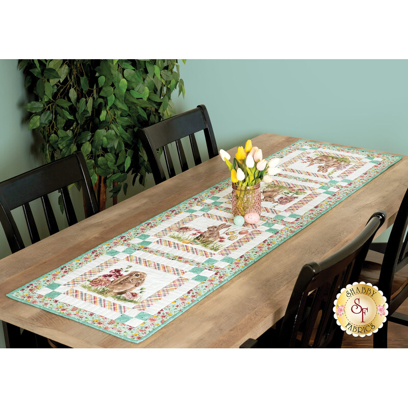 The completed table runner in pink and teal fabrics, set on a wooden table with tulips displayed in a pink glass vase and decorative Easter eggs.