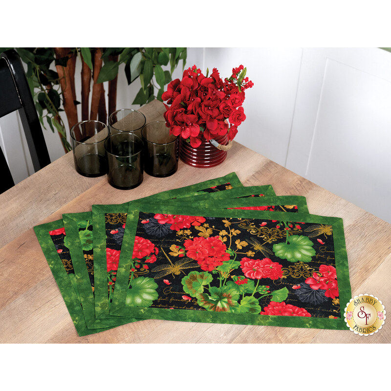 Four floral placemats with red flowers and green leaves sit on a wooden table beside a vase of red flowers.