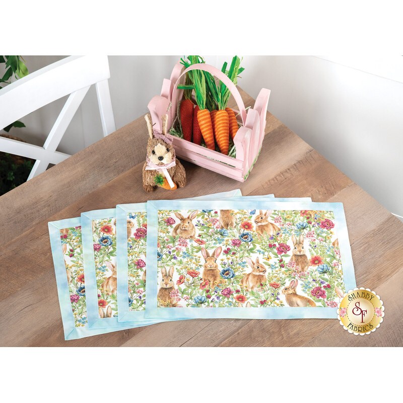 Set of decorative placemats featuring rabbits and flowers, alongside a pink basket with carrots and a bunny figurine on a wooden table.