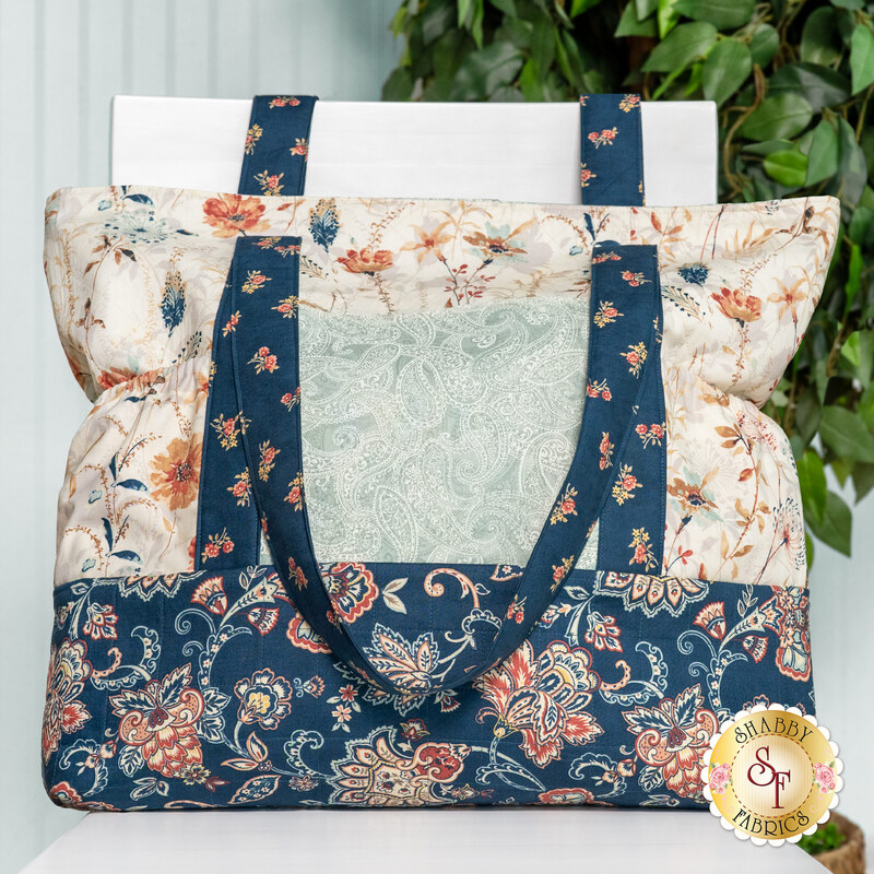 A head-on shot of the completed Carry-It-All Bag, colored in subtle floral fabrics from the Sandalwood collection. The bag is staged on a white chair against a blue wall and leafy tree.