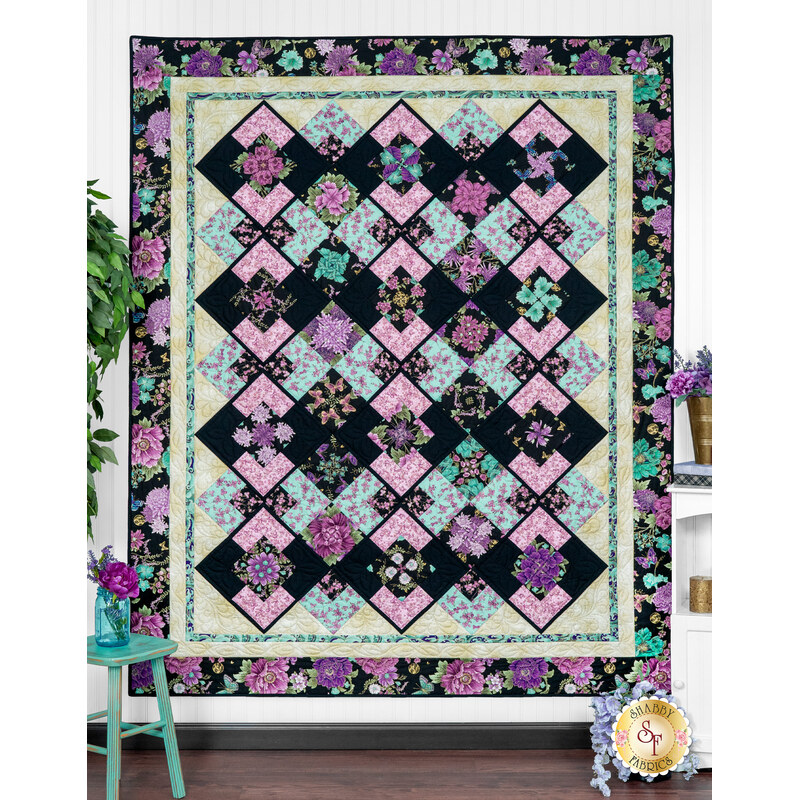 The completed Belle Kaleidoscope quilt colored in purple and aqua floral fabrics from Shizuka, hung on a white paneled wall and bordered by aqua and purple coordinating decor.