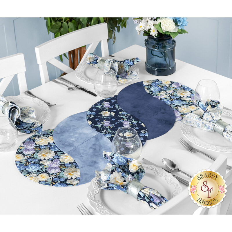 A dining table set with a floral patterned runner, blue napkins, plates, utensils, and a vase of flowers.