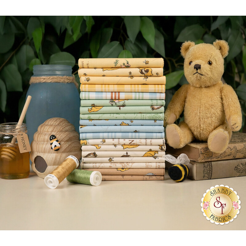 A stack of the fabrics included in the Winnie the Pooh fabric collection, with books and a tree and a teddy bear and bees and a jar and a jar of honey and a beehive and thread.