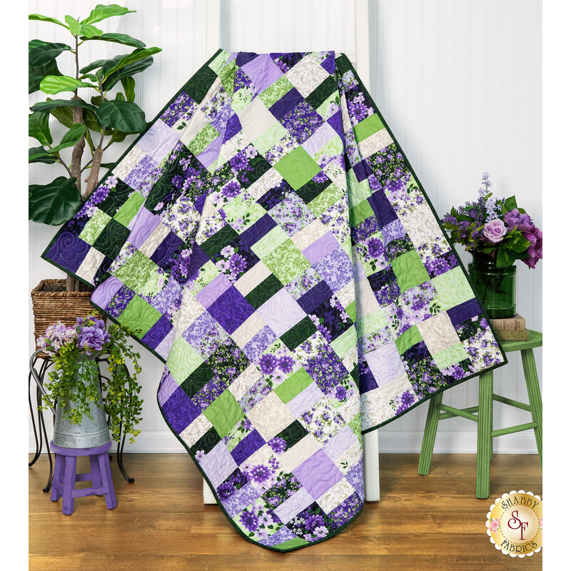 The completed Ten Square Scramble Quilt, colored in bright purple, cream, and green fabrics from the Georgina collection, artfully draped over a white quilt ladder and staged with coordinating greenery and decor.