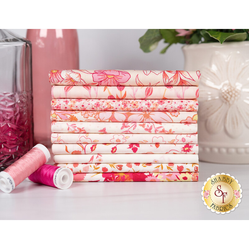 A stack of the fabrics included in the Bloomcore Blush FQ set, with a vase and bottles and thread