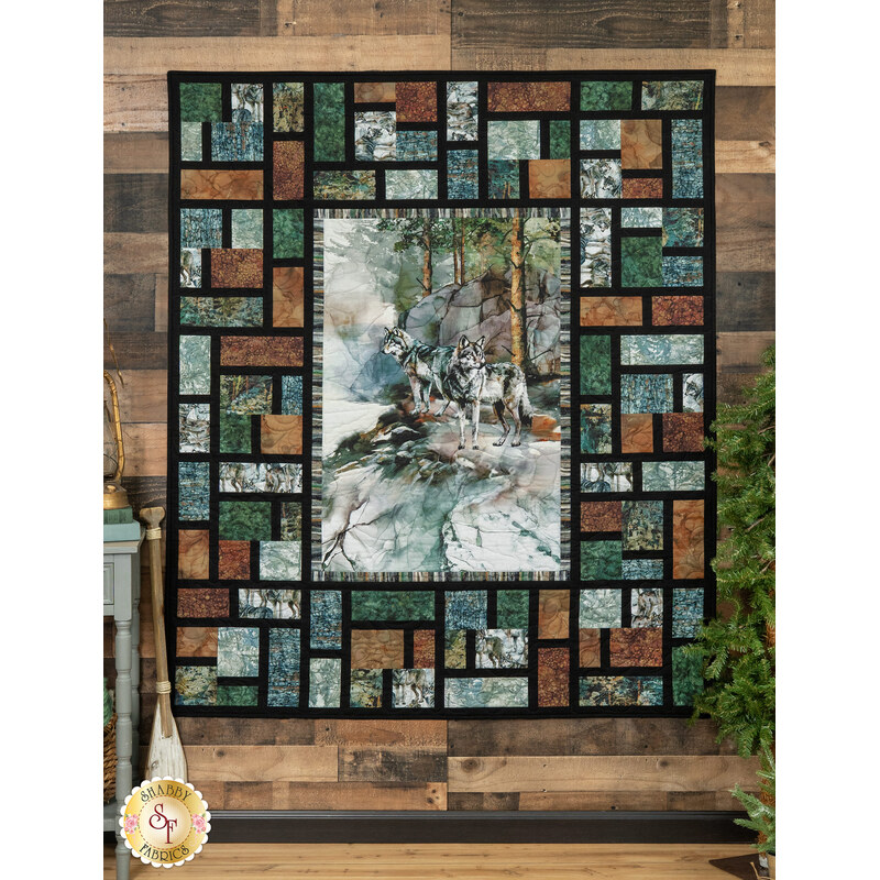 The completed Lone Wolf Stained Glass Panel Quilt, colored in rich green, brown, and blue hues with a watercolor wolf panel centerpiece, hung on a brown wood paneled wall with coordinating decor and greenery.