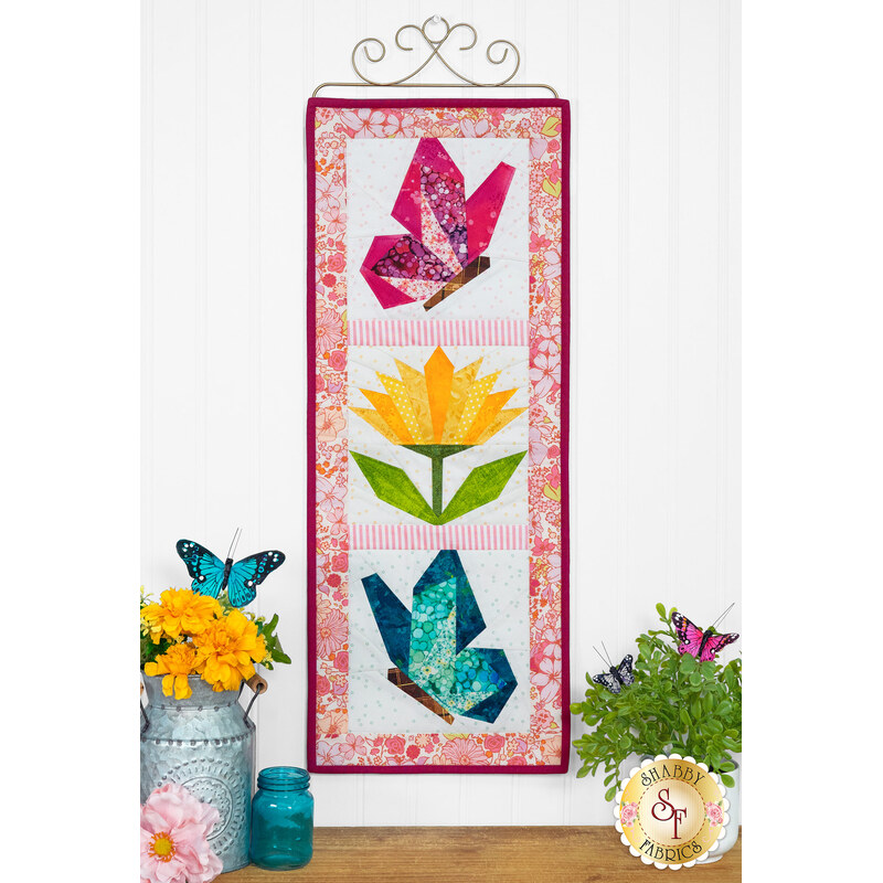 The finished Foundation Paper Piecing Series 3 June project colored in early summer colors, hung on a golden craft hanger on a white paneled wall above coordinating butterfly decor set on a wooden tabletop.