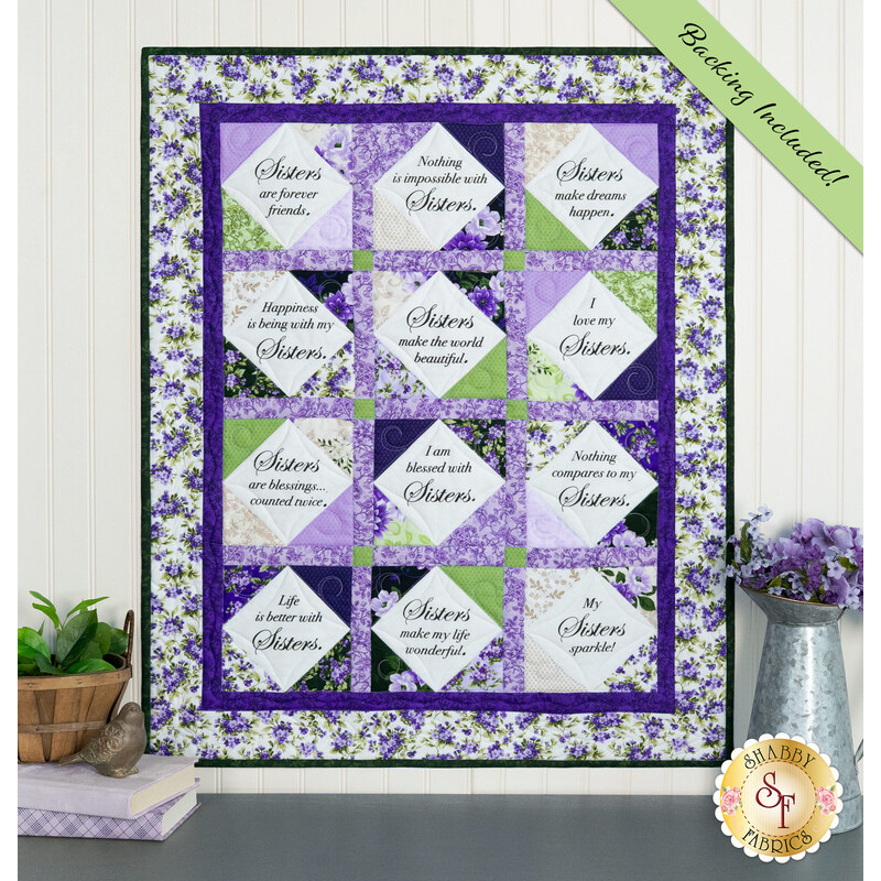 The completed Forever Sisters Charm Quilt colored in vibrant purple, green, and cream fabrics from the Georgina collection, hung on a cream paneled wall above a gray armoire, staged with coordinating decor.