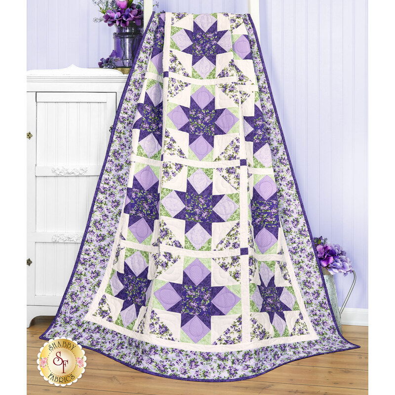 A colorful purple and green quilt with floral and geometric patterns draped over a ladder, staged in front of a light purple wall with coordinating decor and florals.