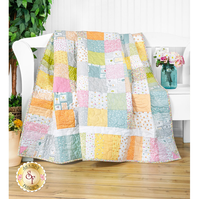 A colorful patchwork quilt draped over a white bench, with a floral vase in the background.