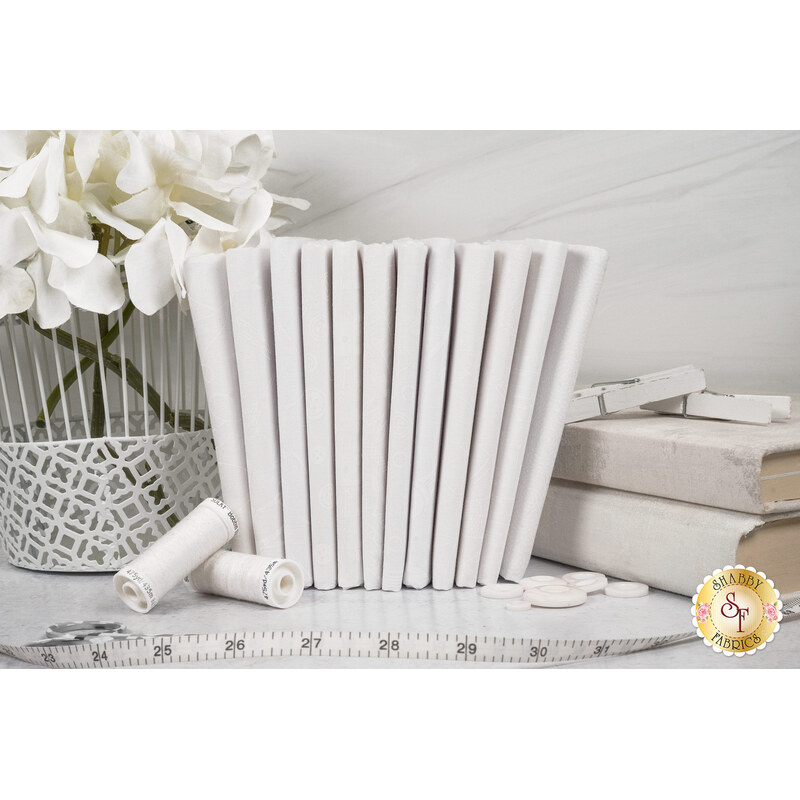 A stack of the white on white fabrics included in the Essentials Sew Happy 2 collection, with books and a basket and thread and buttons and a measuring tape and flowers.