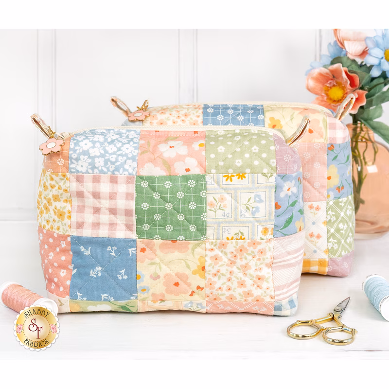 The two completed Mini-Charm Bags, colored in gentle pastels from the Cottage Charm collection by Moda, staged on a white table with coordinating notions and flowers.