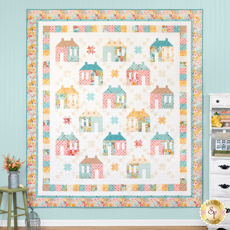 The completed Maisons de Patchwork Quilt colored in bright yellow, pink, and blue fabrics, hung on a blue paneled wall and bordered by coordinating summer decor.