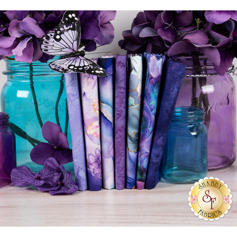 A stack of the fabrics included in the Flourish Amethyst FQ set, with jars and flowers and butterflies and thread.