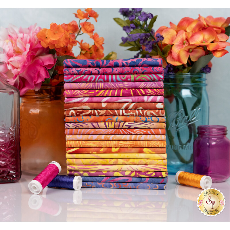 A stack of the batik fabrics included in the Sunset collection, with mason jars and flowers and thread.