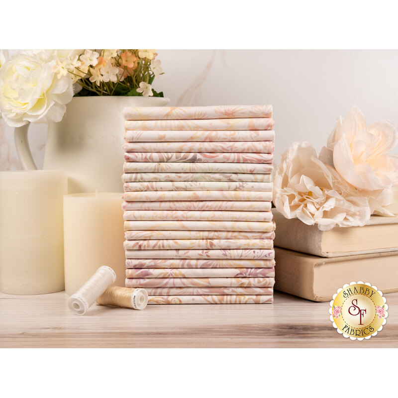 A stack of the fabrics included in the Quiet Charm collection, with candles and books and a pitcher and flowers and thread.
