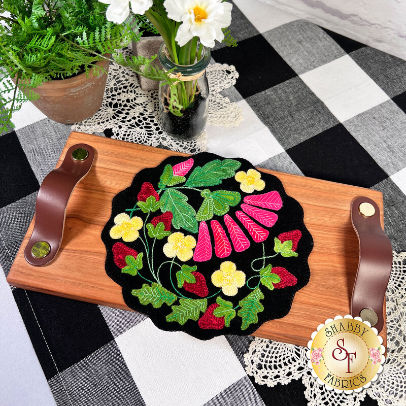 A top down shot of the completed Strawberry Zinger wool mat kit in black and bright colors, staged on a cutting board on a table with coordinating decor.