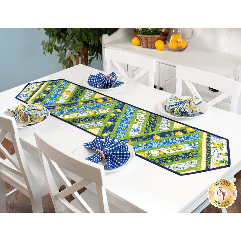 The completed Greased Lightning Table Runner in green, blue, and yellow fabrics from the Limoncello collection, staged on a white table with coordinating cloth napkins and summer decor.