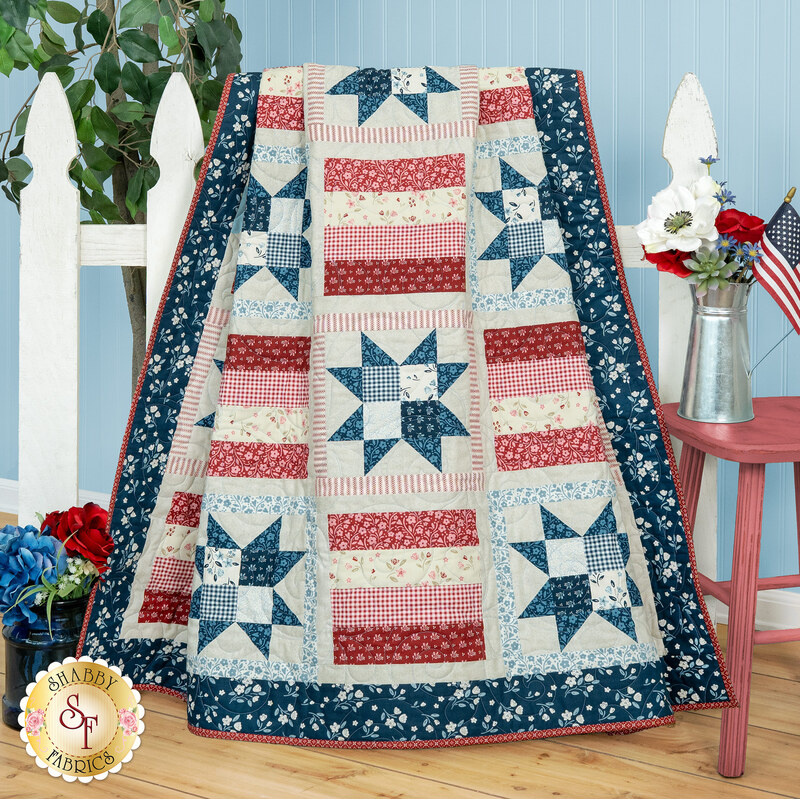 A patriotic quilt with star patterns in red, blue, and cream draped over a white picket fence next to flowers and coordinating decor. 