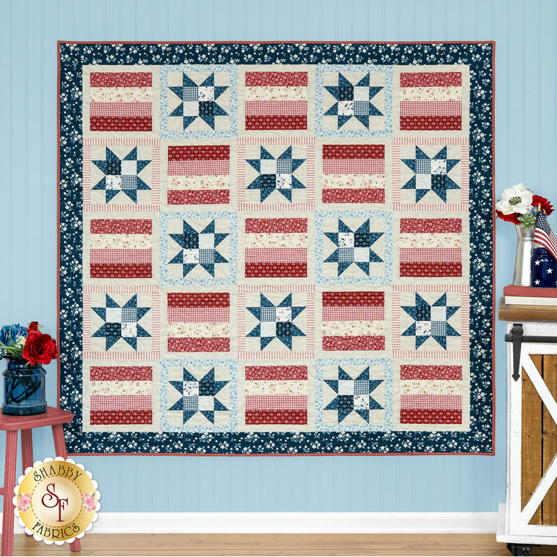 A patriotic quilt featuring star and striped patterns in red, white, and blue, displayed against a blue wall next to coordinating decor. 