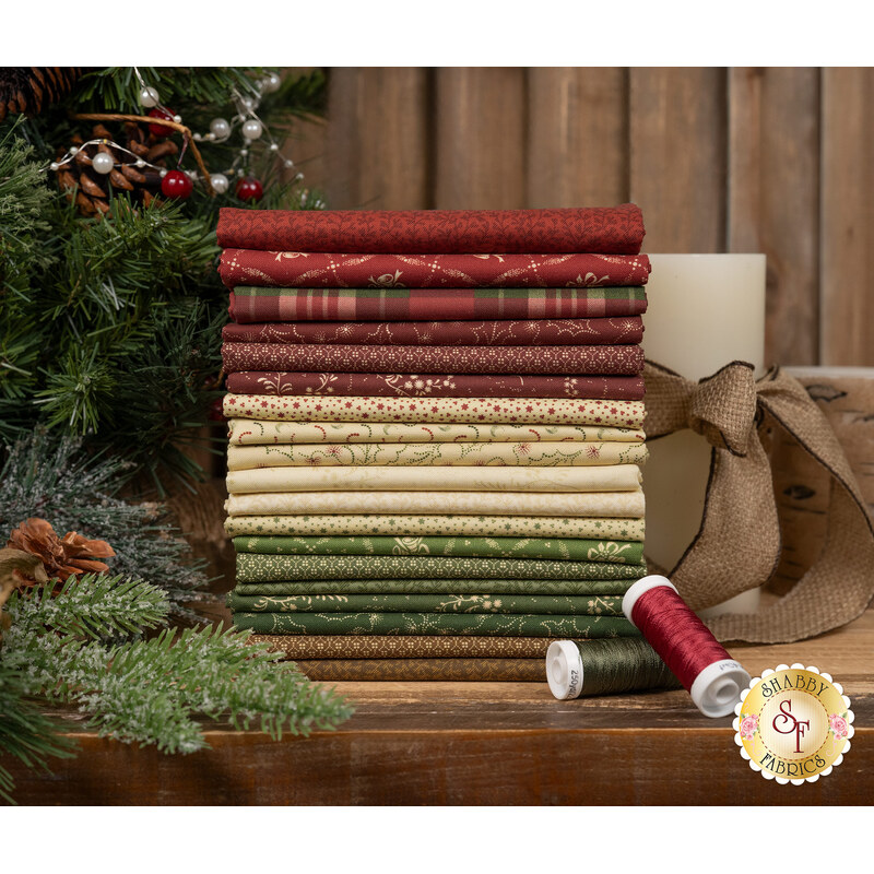 A stack of red, brown, tan, and green Christmas fabrics included in the Cozy Christmas FQ Set with pine branches and a candle and ribbon and thread.