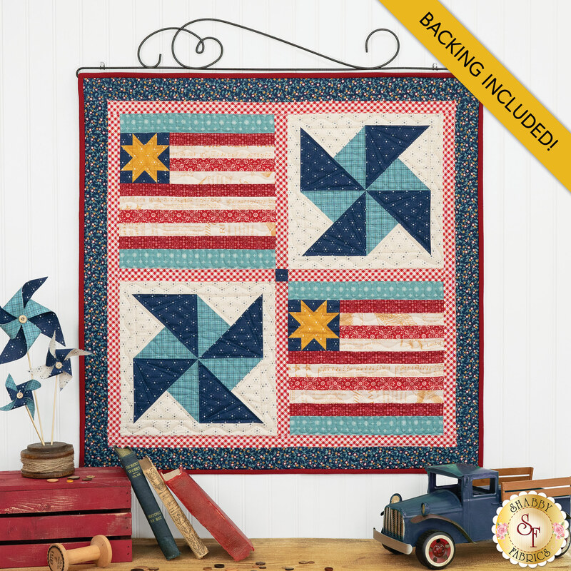 The completed July Pieceful Patchwork wall hanging, colored in cream, red, blue, and teal fabrics from the Americana collection by Lori Holt for Riley Blake Designs. The project is hung on a white paneled wall and staged with coordinating vintage americana decor.