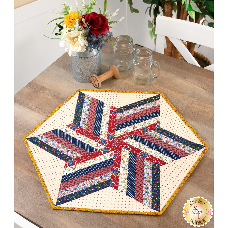The completed 60 Degree Diamond Table Topper Kit colored in red, cream, blue, and yellow fabrics from the Old Glory collection by Henry Glass fabrics, staged on a rustic table with coordinating decor.