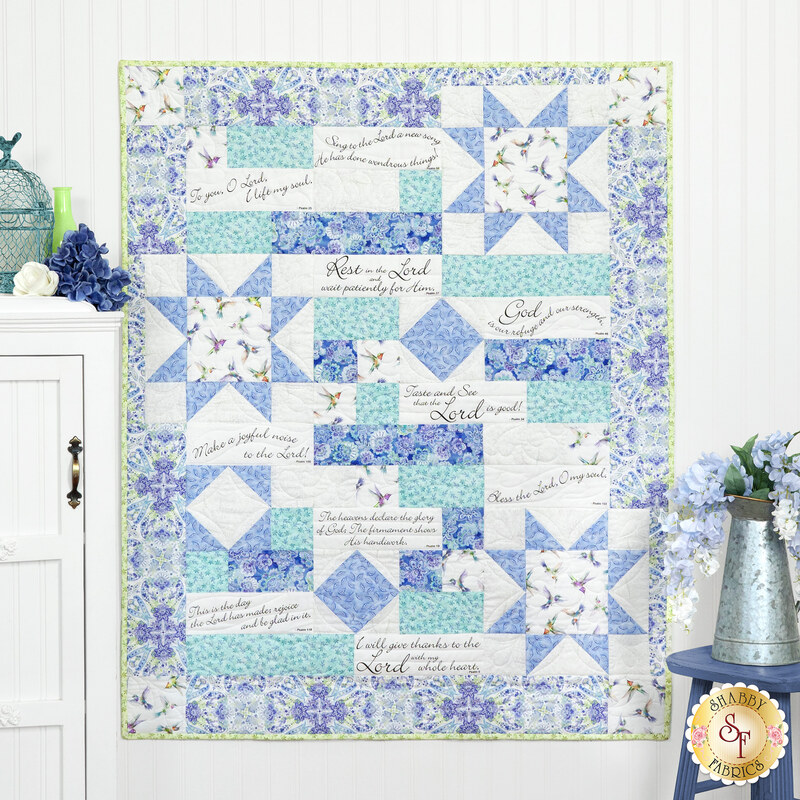 The completed Comfort of Psalms Quilt colored in blue, aqua, white, green, and purple fabrics from the Daydreams collection, staged on a white paneled wall with coordinating decor and furniture.