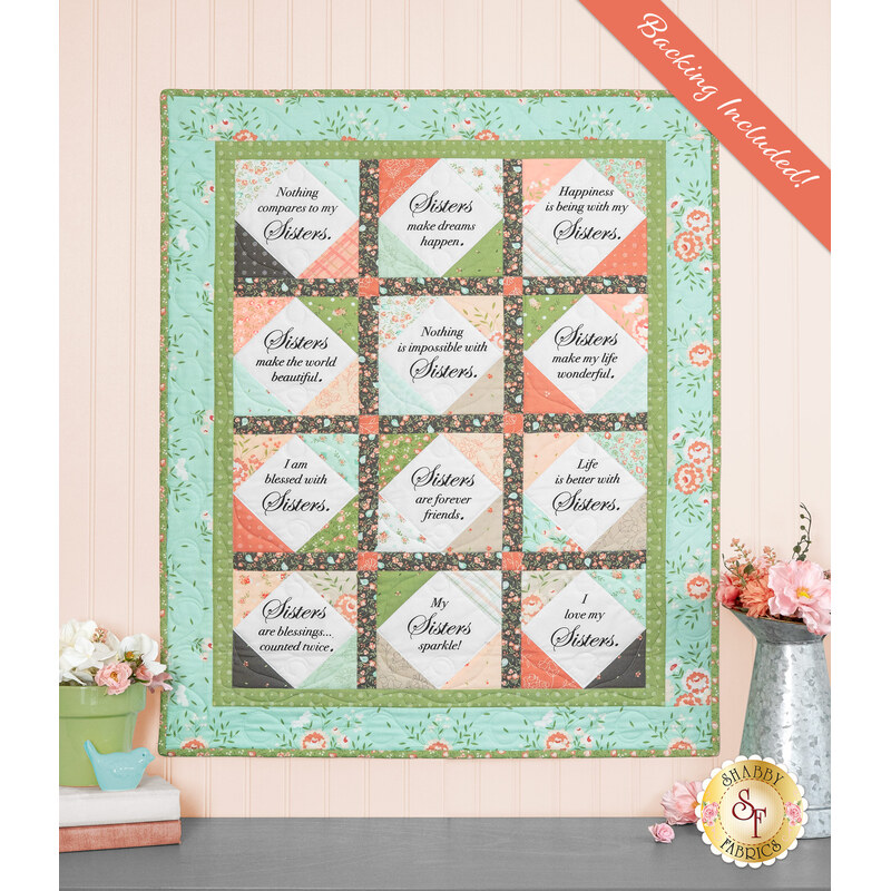 The completed Forever Sisters Charm Quilt colored in vibrant aqua, green, charcoal, and pink fabrics from the Abloom collection, hung on a cream paneled wall above a gray armoire, staged with coordinating decor.