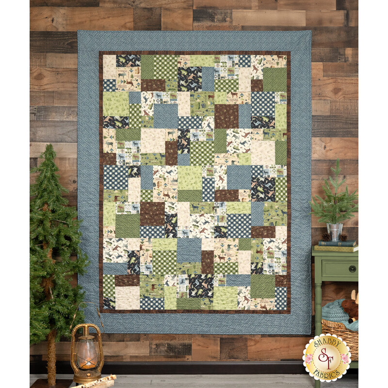 The Easy as ABC and 123 Quilt - Lodge Life quilt featuring a patchwork design in tones from nature like blue, green, and brown, hanging from a wood-paneled wall next to green trees and coordinating decor.