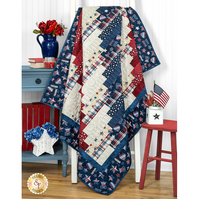 The Standing Strong quilt draped over a chair, with red, white, and blue patterns, alongside flowers and decor.