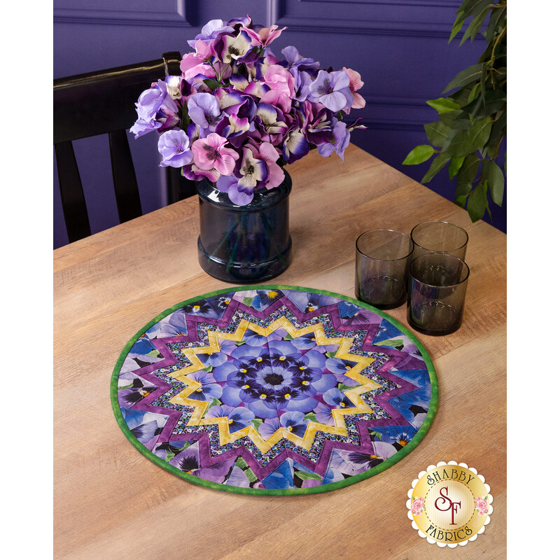 The completed Point of View Kaleidoscope Folded Star Table Topper colored in vibrant purple fabrics from the Pansy Perfection collection, staged on a wooden table with coordinating flowers and glass ware.