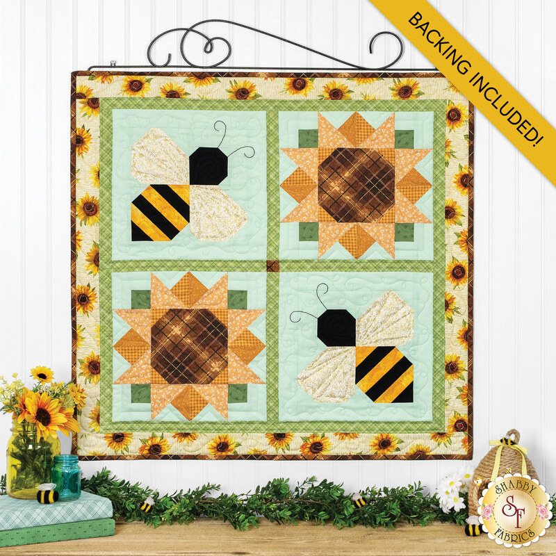 The completed Pieceful Patchwork - August wallhanging, a sunflower and bee-themed project colored in light blue, gren, cream, and golden yellow fabrics. The project is hung on a white paneled wall and staged with coordinating decor.