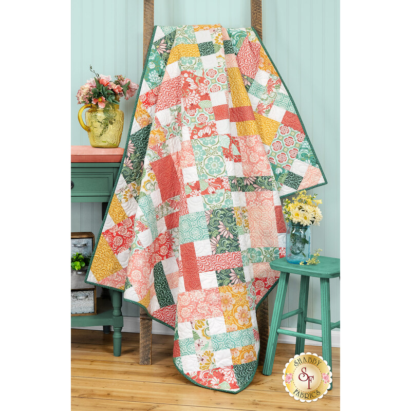 The completed Picnic Quilt colored in vibrant, peach, gold, aqua, and teal fabrics from the Tango collection by Moda Fabrics. The project is hung on a rustic wooden quilt ladder and staged with coordinating flowers and furniture.