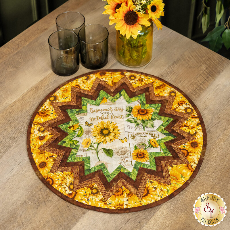 The completed Point of View Folded Star Table Topper colored in bright yellow, brown, green, and cream fabrics from the Sunflower Farm collection. The project is staged on a rustic wood table with coordinating glass ware and flowers.
