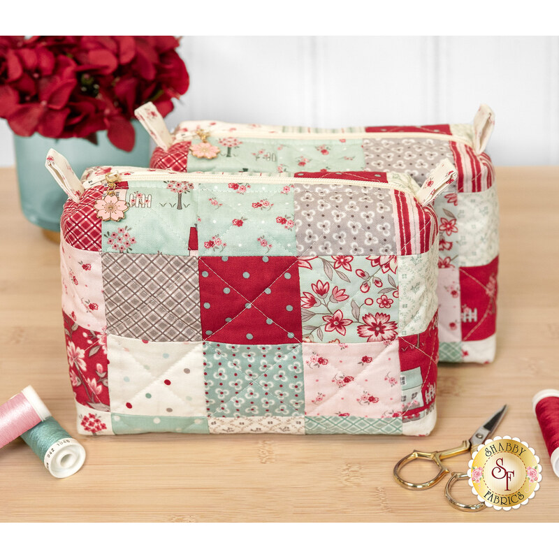 The two completed Mini-Charm Bags, colored in the rich hues from the My Summer House collection by Moda, staged on a wooden table with coordinating notions and flowers.
