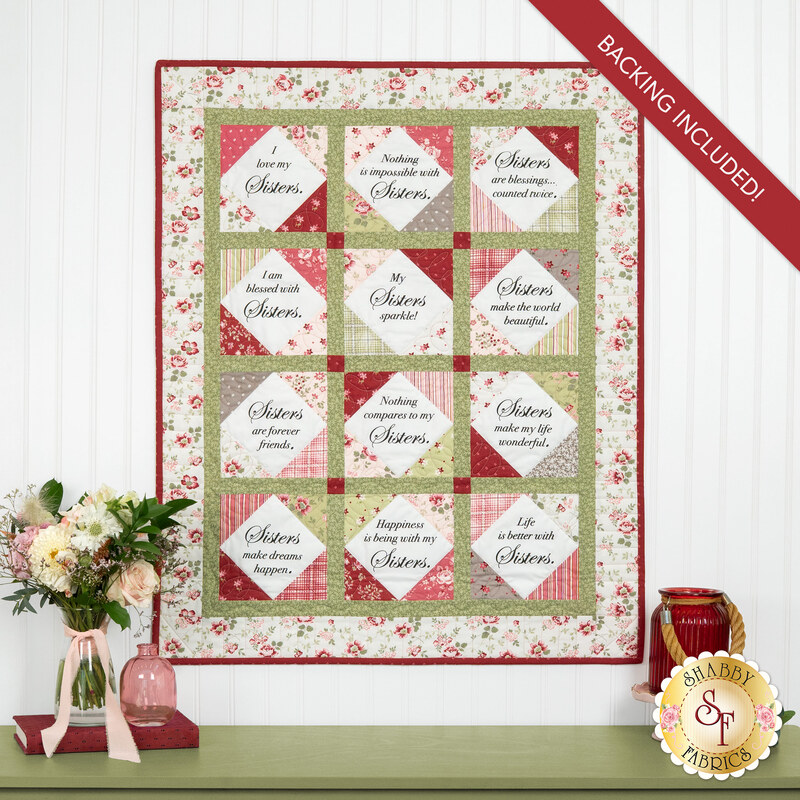 The completed Forever Sisters Charm Quilt colored in rich red, pink, and green fabrics from the A Very Sweet Christmas collection, hung on a white paneled wall above a green armoire, staged with coordinating décor.