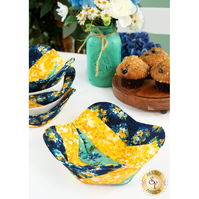 A completed bowl cozy colored in navy, yellow, and teal, staged on a white table with a stack of other bowl cozies, coordinating florals, and blueberry muffins on a wooden platter.