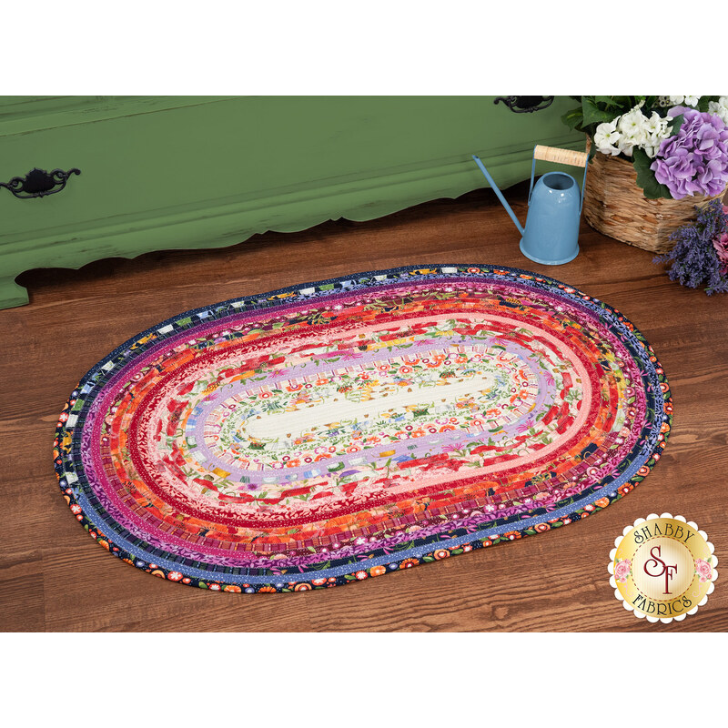 The completed Jelly Roll Rug colored in vibrant red, pink, purple, and white fabrics from the Superbloom collection by Moda Fabrics, staged on a wooden floor beside a green armoire and coordinating decor.