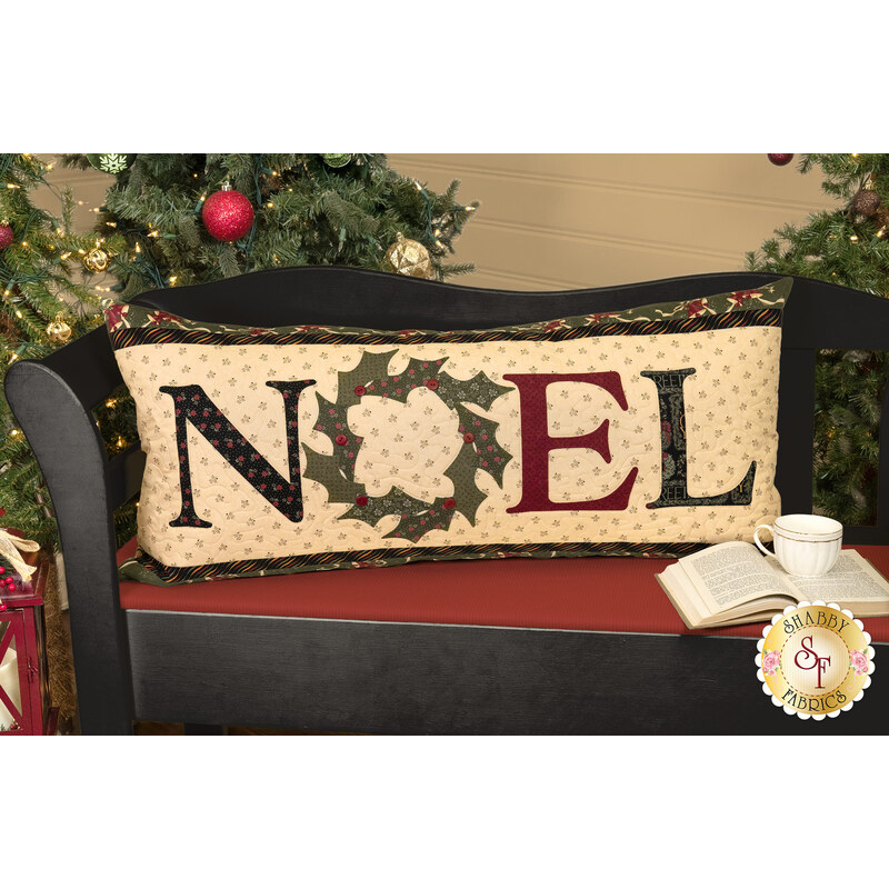 The completed pillow colored in festive A Vintage Christmas fabrics, staged on a black and red bench with a cup of tea and a book, in front of a Christmas tree set.