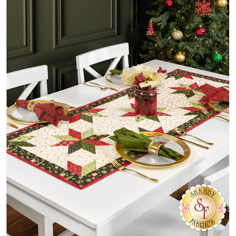 The completed table runner colored in cream, red, and green floral Christmas fabrics from the Glisten collection, staged on a white table with coordinating gold-rimmed plates, cloth napkins, florals, and a decorated Christmas tree.
