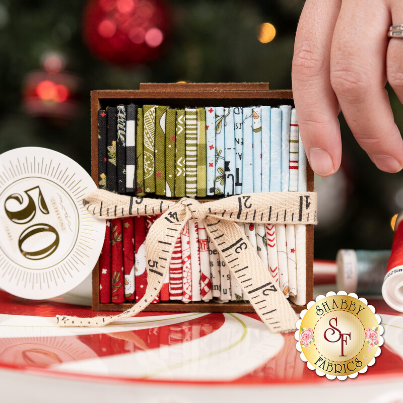 A wooden box of colorful fabric rolls, tied with a measuring tape ribbon, on a festive table. A model's hand peeks into frame from the right side.