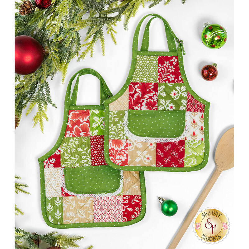 The two completed hot pads colored in bright green, red, and cream fabrics, from the Buon Natale collection, staged on a clean white background with coordinating holiday decor and a wooden spatula.