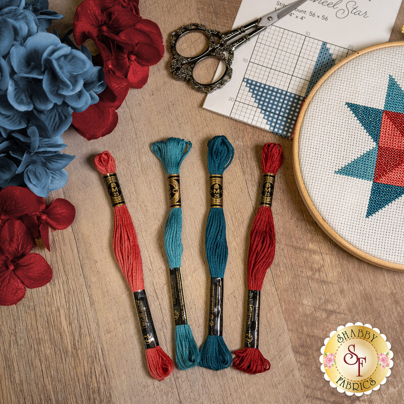 A shot of four skeins of embroidery floss in red, burgundy, teal and blue, fanned out on a wooden table with an embroidery project in its wooden hoop peeking into the top right corner next to red and blue flowers.