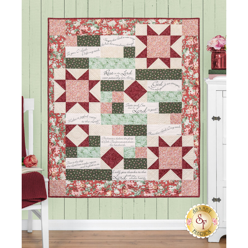 The completed Comfort of Psalms Quilt Kit, colored in red, green, cream, and pink fabrics from the Regal Rose collection, hung on a green-paneled wall and staged with coordinating furniture and décor.