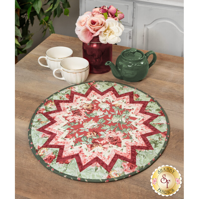 The completed Point of View Folded Star Table Topper colored in mint green, red, and pink floral fabrics from the Regal Rose collection. The topper is staged on a wooden table with coordinating red and green tableware and flowers.