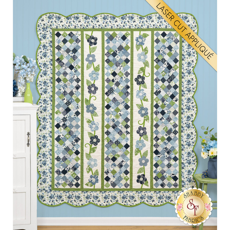 The completed Flower Girl quilt colored in green, cream, blue, and navy from the Nantucket Summer collection, hung on a blue-paneled wall with coordinating floral décor on either side of the quilt.