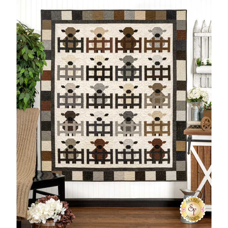 The completed Charming Lambs quilt colored in earthy neutrals from the Woolies Flannel collection, depicting five rows of four lambs behind little fence doors, hung on a white paneled wall and staged with coordinating farmhouse decor.