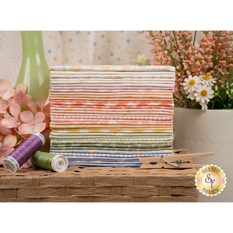 A stack of the fabrics included in the Pastel Prairie set, with a vase of flowers and clothes pins and thread.